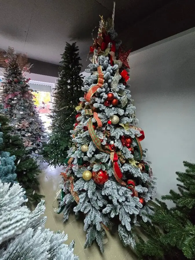 Brad artificial înalt, împodobit cu lumini calde și decorațiuni roșii, lângă o fereastră.